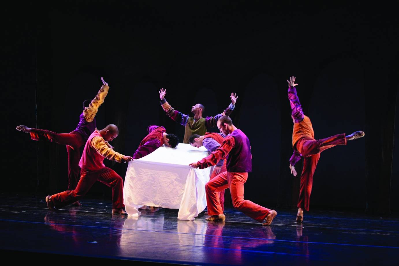 Male dancers dressed in reds, yellows and purples cluster around a table with a white tablecloth